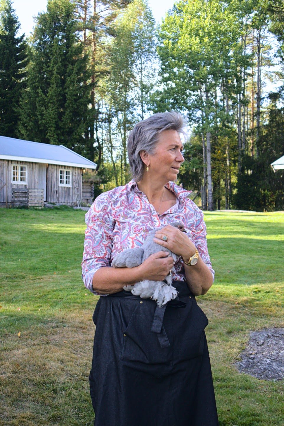 Marianne i mønstret skjorte og forkle holder en grå høne på armen ute på tunet, med et gråværet uthus og høye trær bak
