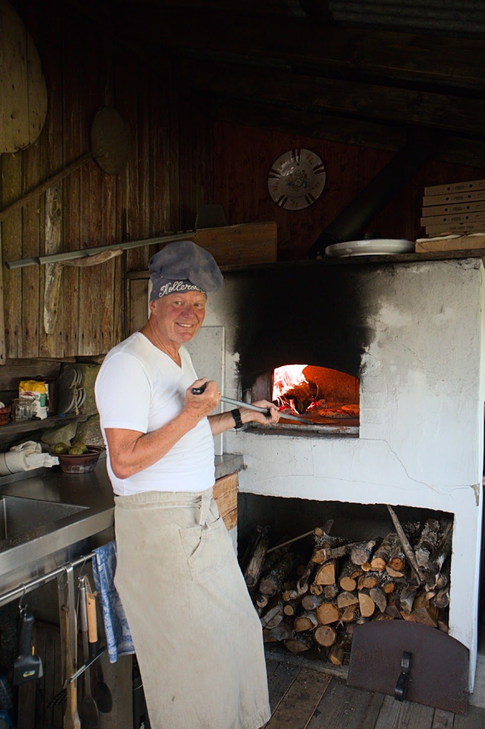 Lars i kokkelue med «Kollerøs» smiler ved den hvite vedovnen i utekjøkkenet, med pizza i ovnen og ved stablet under