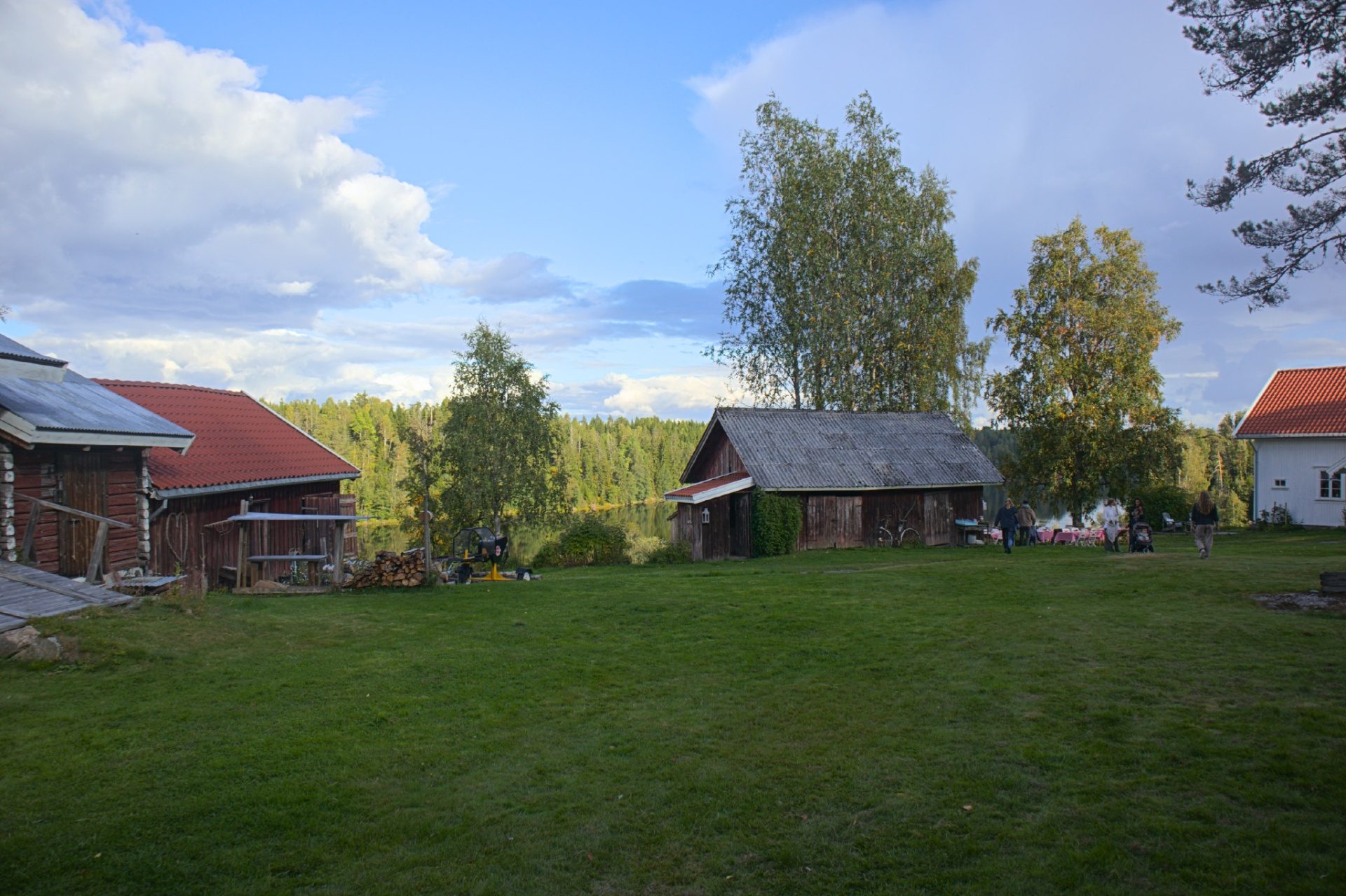 Det store, grønne tunet med gamle røde og grå tømmerbygninger, bjørker og innsjøen bak, og gjester på vei mot bordene