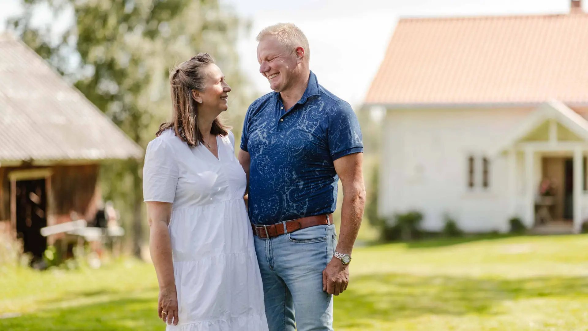 Marianne og Lars smiler til hverandre på plenen, med det hvite hovedhuset og et gammelt uthus i bakgrunnen