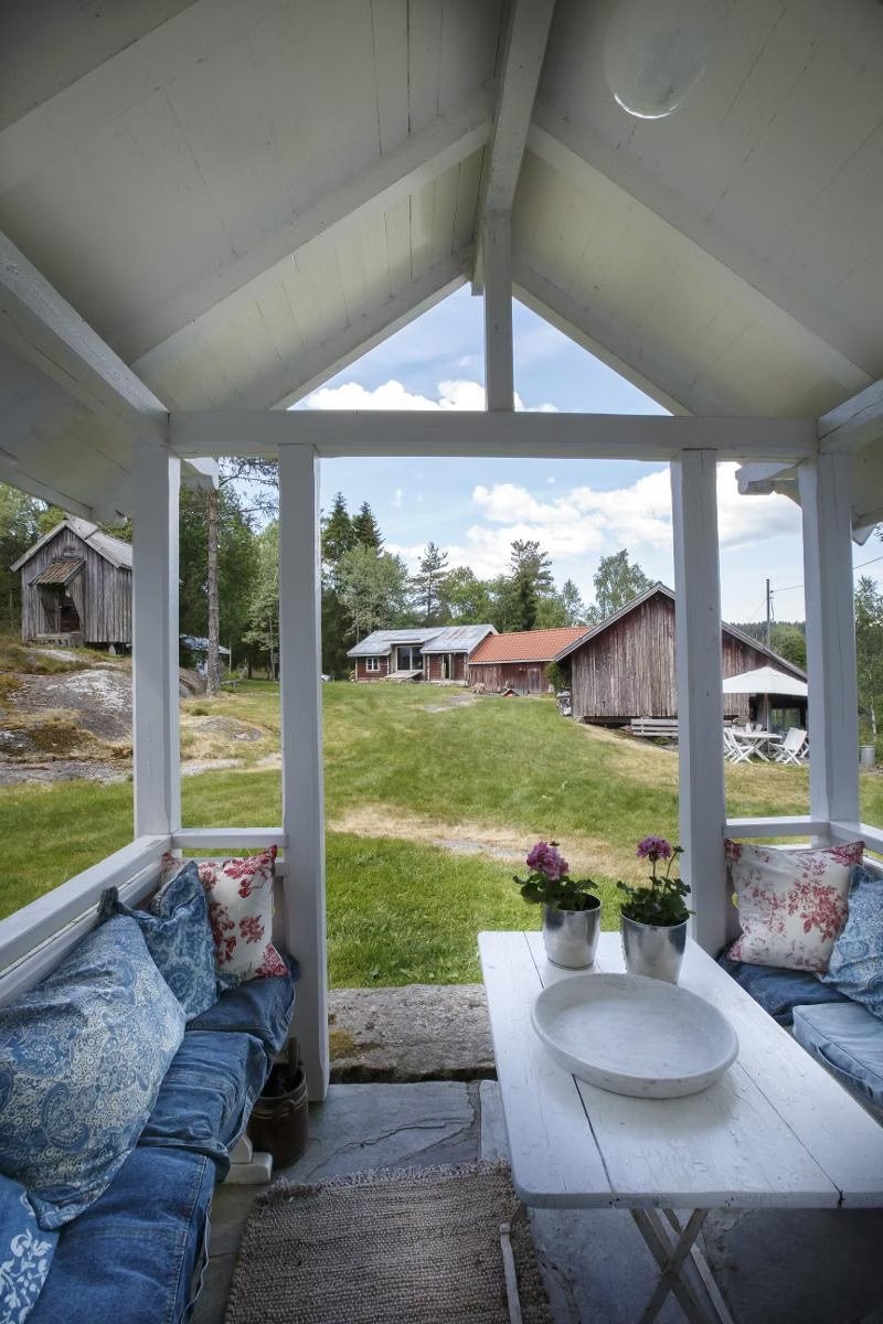 Utsikt fra et hvitt, åpent lysthus med puter og pelargonier på bordet, ut over plenen mot de gamle bygningene