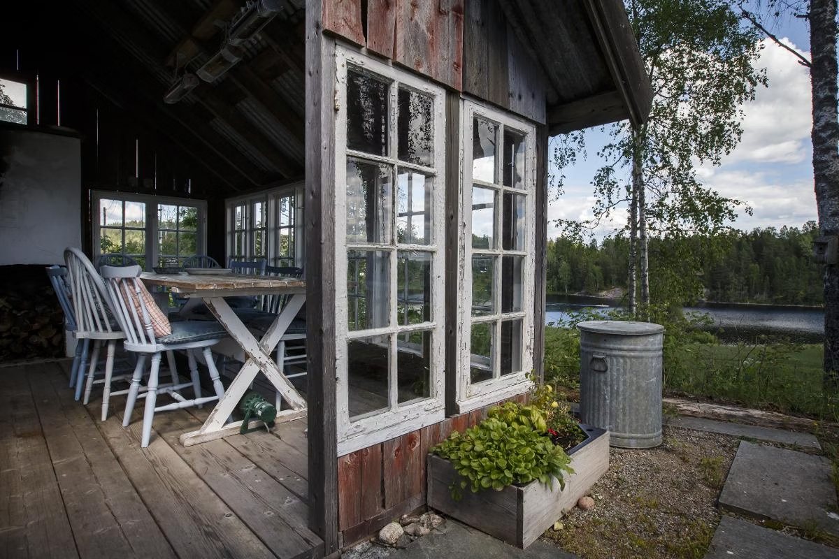 Utekjøkken under tak med langbord, hvite pinnestoler og gamle sprossevinduer, med utsikt mellom bjørkene mot vannet