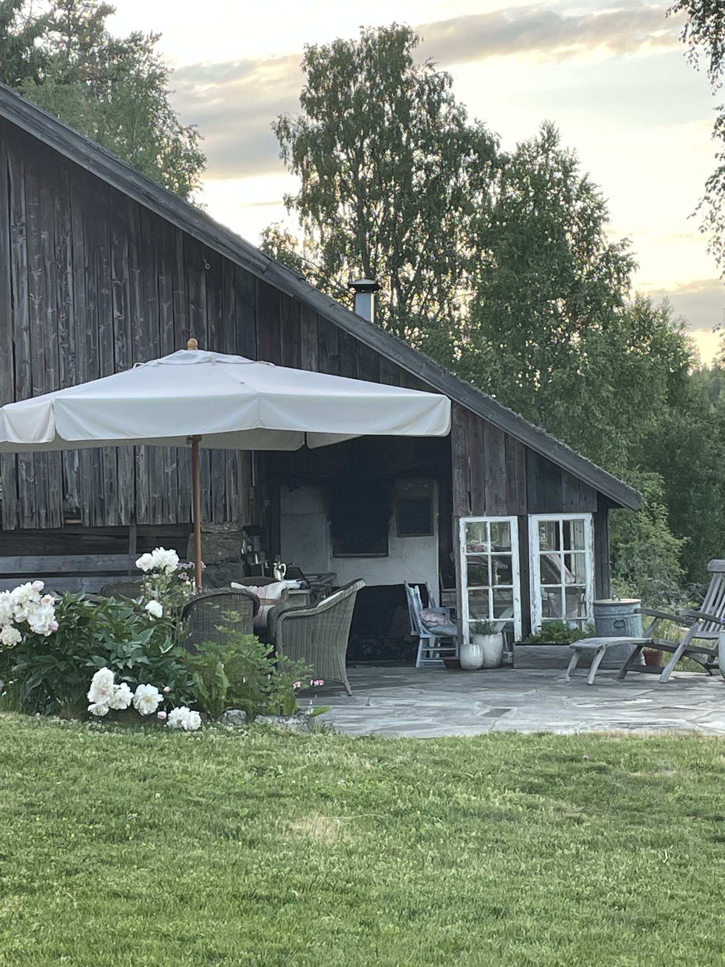 Utekjøkkenet i den gamle tømmerbygningen en sommerkveld, med hvit parasoll, kurvstoler, hvite peoner og gamle sprossevinduer