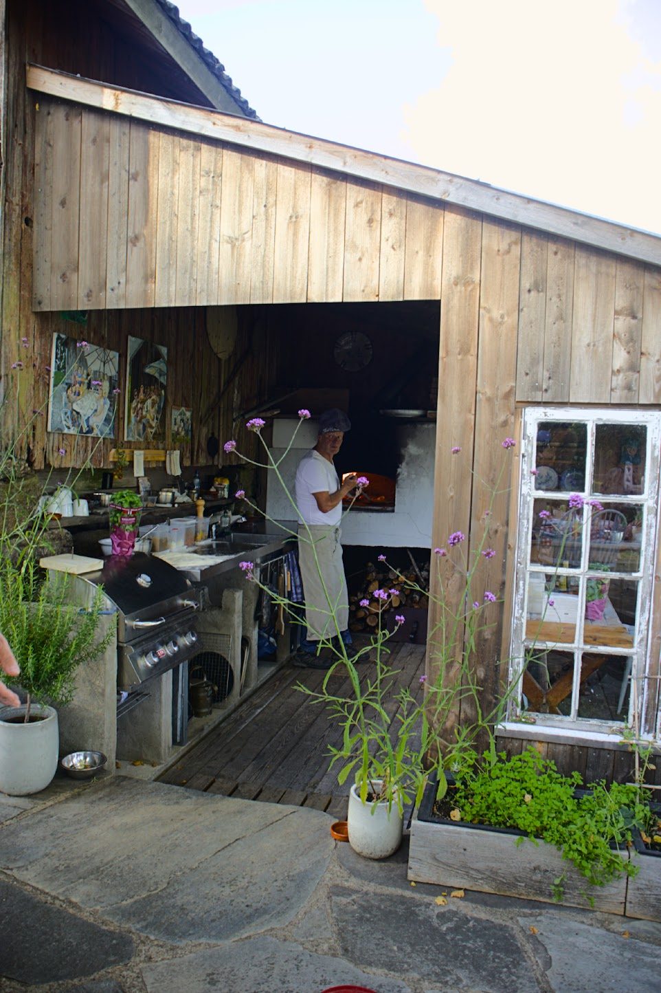 Utekjøkkenet sett fra hellene utenfor, der Lars i forkle og kokkelue fyrer i den hvite vedovnen, med blomster og et gammelt sprossevindu i forgrunnen