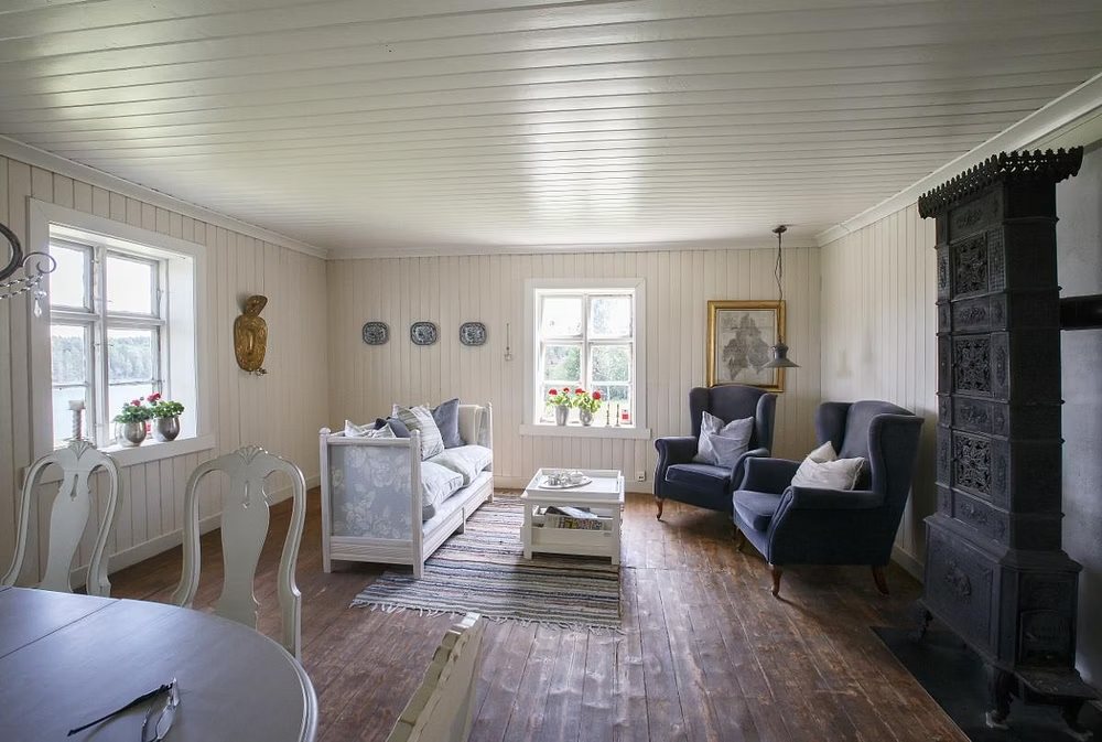 Stue i hovedhuset med hvite panelvegger, gamle furugulv, ørelappstoler, en gammel sofa og en svart støpejernsovn