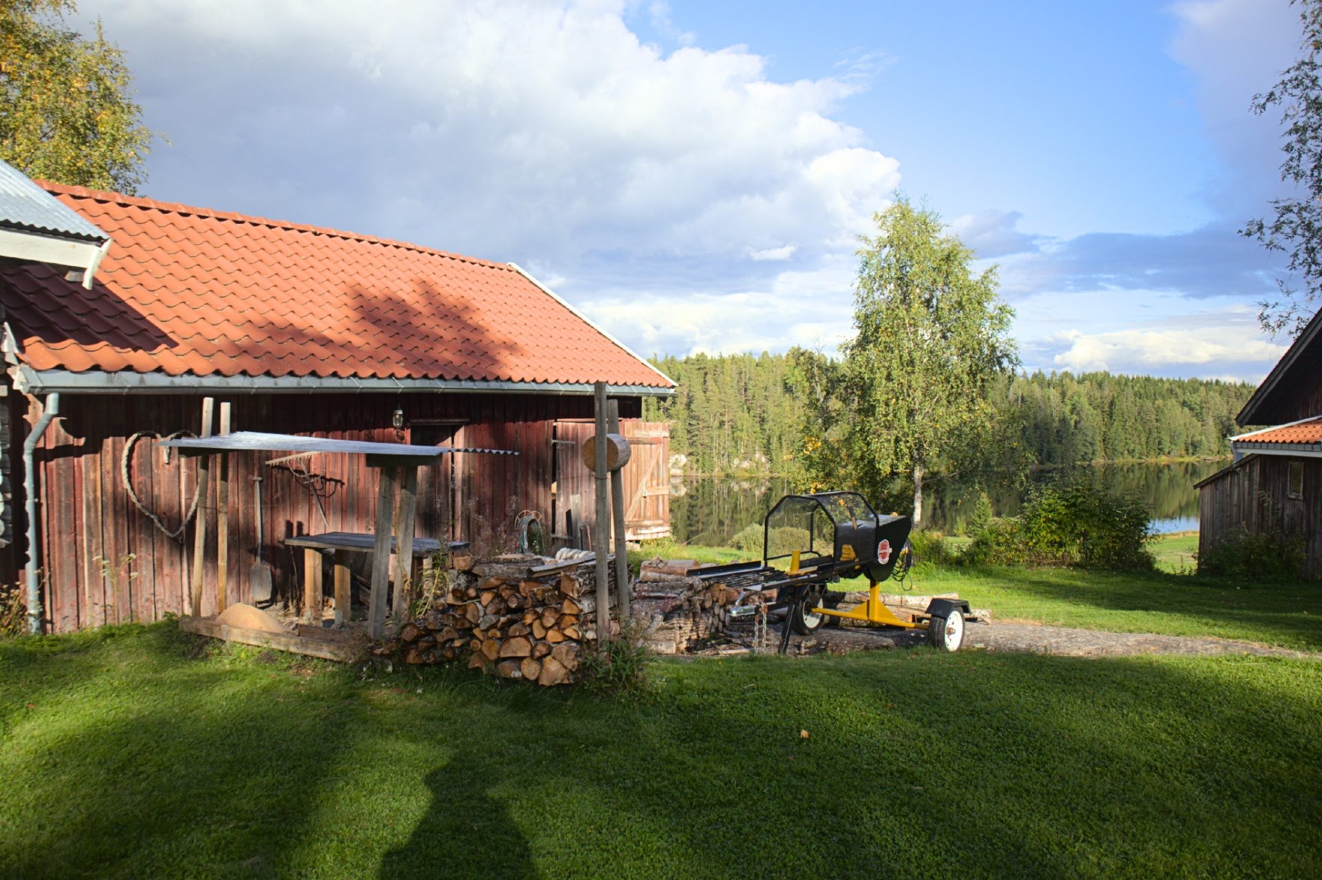 Stablet ved og en vedkløyver foran et rødt tømmerhus med tegltak, med bjørk og innsjøen i bakgrunnen